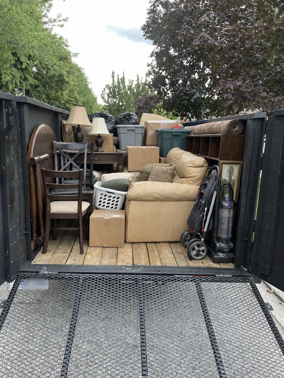 A loaded utility trailer holding an armchair, wooden chairs, boxes, lamps and other household items