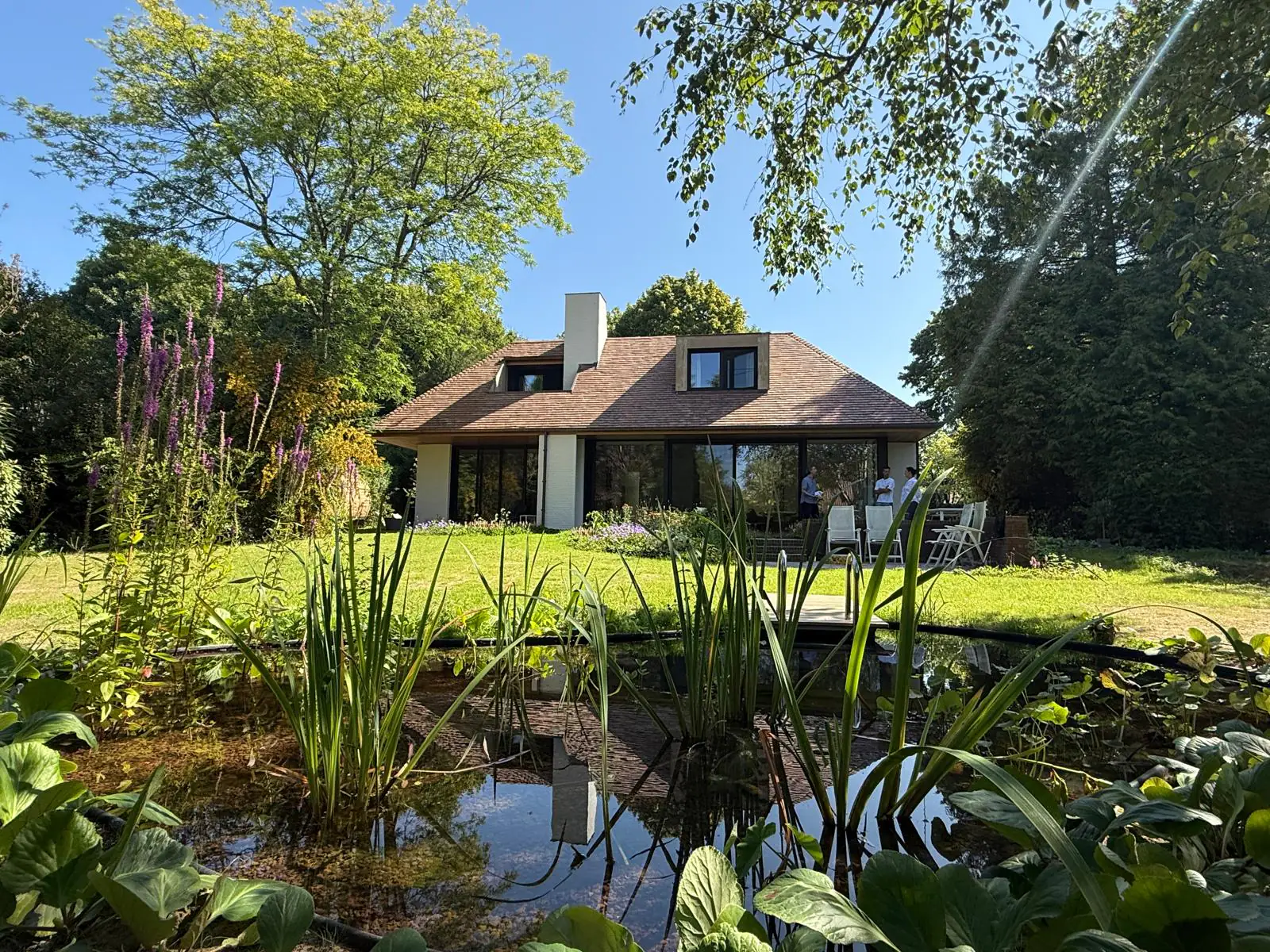 Het huis van B&Biotoop achter het gazon en de spiegelende tuinvijver in de ochtendzon